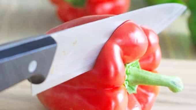 A red bell pepper on its side with a knife cutting off the stem end.