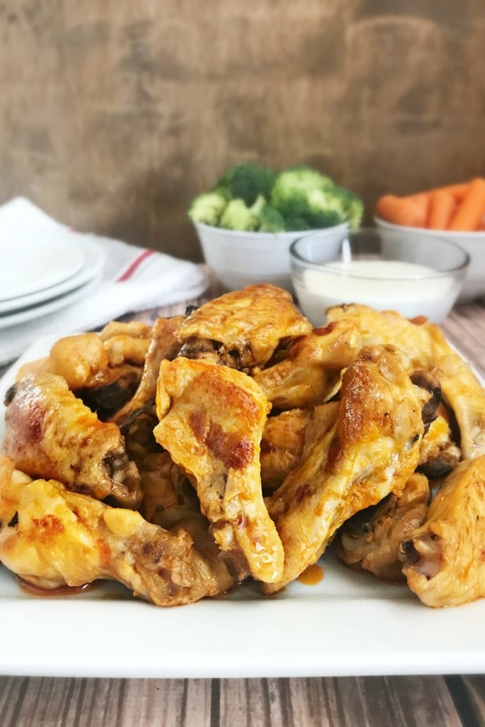 Plate full of golden chicken wings. In the background is a glass bowl with a white dipping sauce, and two white bowls - one filled with broccoli florets and one with baby carrots. Next to the bowls is a white dishtowel with a red stripe and a stack two round plates.