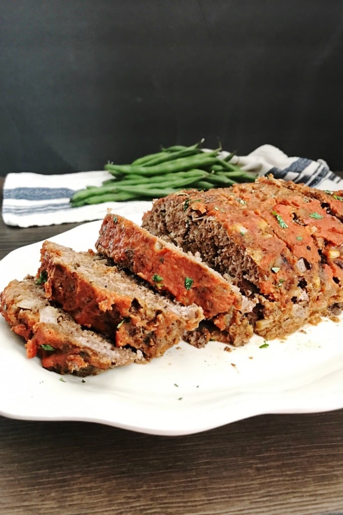 Slow Cooker Meatloaf Whole meatloaf, sliced, topped with red sauce and garnished with diced parsley on a white serving platter. In the background is a dishtowel with a pile whole raw green beans.