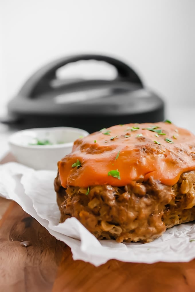 Meatloaf sitting on a piece of white parchment paper on a wooden cutting board topped with a red sauce and garnished with chopped parsley. In the background is a small white bowl with chopped parsley and behind that is the lid of any Instant Pot.