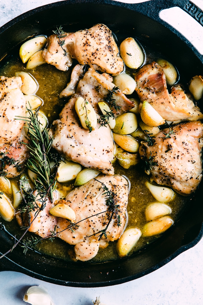 Black cast iron skillet filled with roasted chicken thighs and garlic cloves with fresh thyme and fresh rosemary sprigs. 