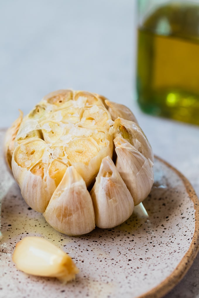 Whole clove of garlic with the top chopped off, roasted to golden and sprinkled with flakey sea salt sitting on small speckled white and brown late with an unpeeled clove of garlic on it as well. In the background you can see a bottle of olive oil.