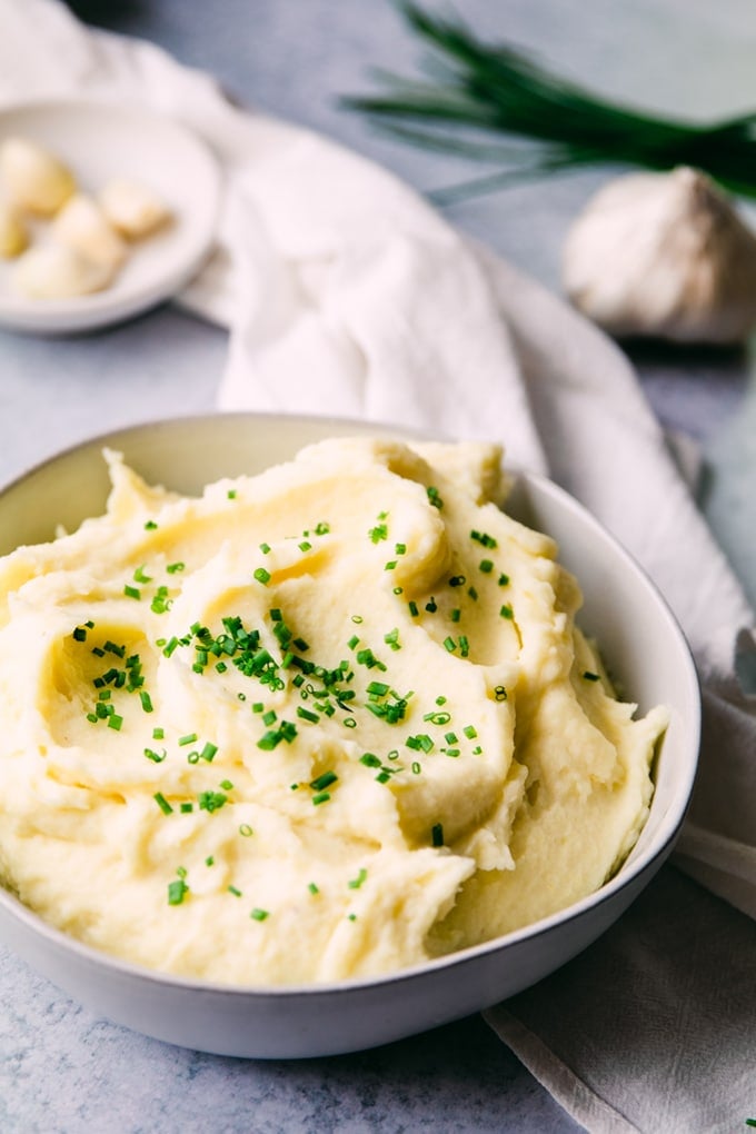 Bowl of mashed potatoes topped with chopped chives. In the background is a whole head of garlic and some peeled garlic cloves in a small white dish.