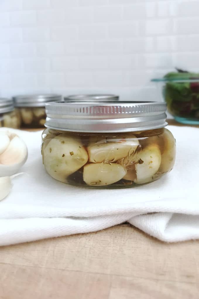 Short mason jar filled with pickled garlic. In the background you can see other short mason jars filled with garlic plus a larger glass bowl filled with greens. To the left are some unpeeled garlic cloves.