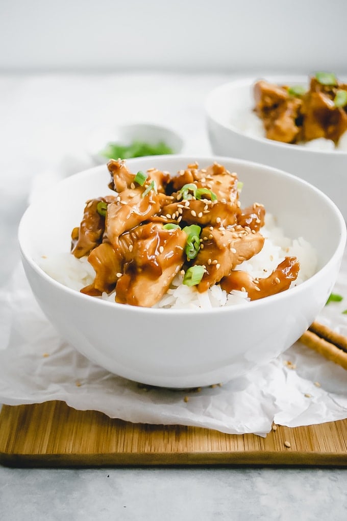 Chicken Garlic Chicken over rice, topped with sesame seeds and scallions, all in a deep white bowl.