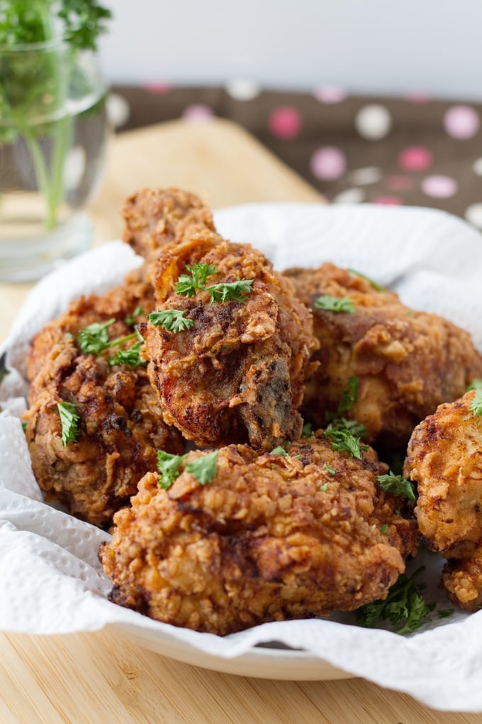 Pieces of fried chicken in a paper towel lined bowl on a light wood cutting board with a some parsley sitting in water in the background.