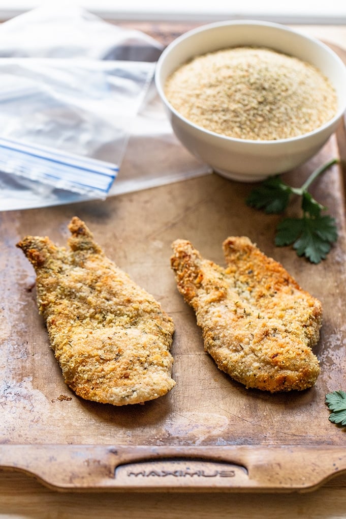 Two fillets of chicken that has been breaded and baked to a golden brown sitting on a wooden cutting board. It is garnished with some parsley sprigs. There's a white bowl of breadcrumbs sitting on the cutting board as well as some empty zip lock bags.