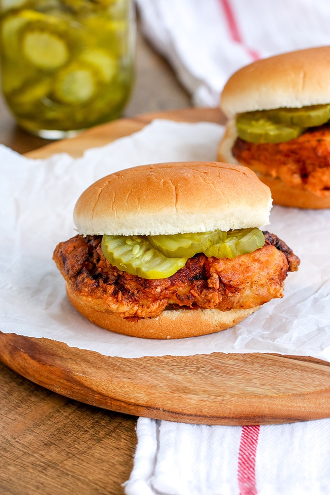 Two fried chicken sandwiches with sliced pickles on them sitting on some white paper on a round wooden cutting board with a jar of sliced pickles in the background.