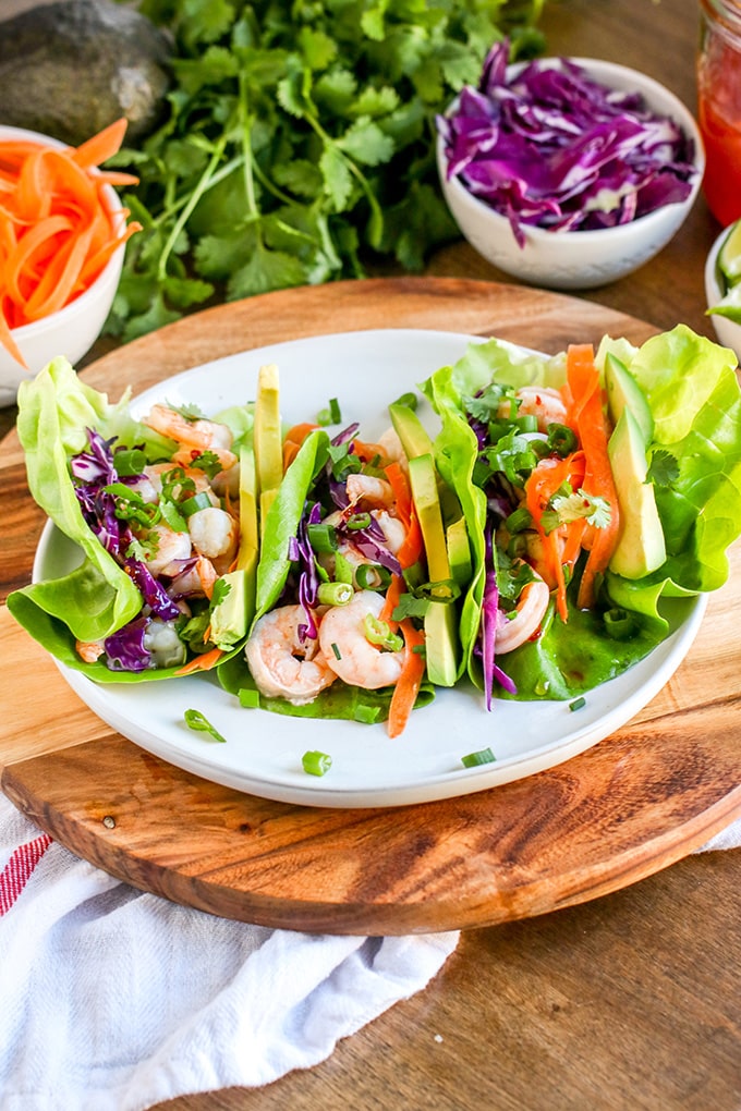 Lettuce wraps on a round white plate filled with red cabbage, shrimp, sliced avocado, shredded carrots, chopped cilantro and chopped green onions. The plate is sitting on a round wood board, and is surrounded by bowls filled with chopped red cabbage, carrot ribbons, and a bunch of cilantro.