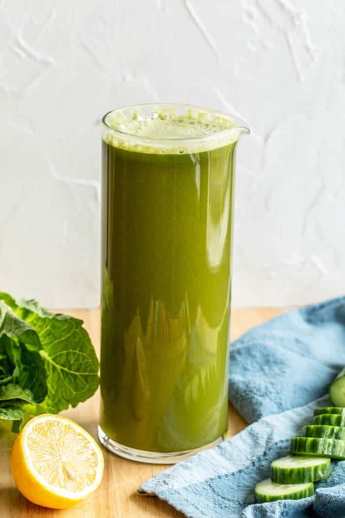 Glass pitcher filled to the brim with green juice. Sitting on a wood cutting board with a half a lemon, some sliced cucumber and a blue dish towel.