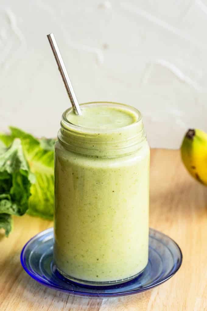 Green smoothie in a glass jar sitting on a small blue plate with a metal straw sticking out of it. In the background is some romaine lettuce and a banana.