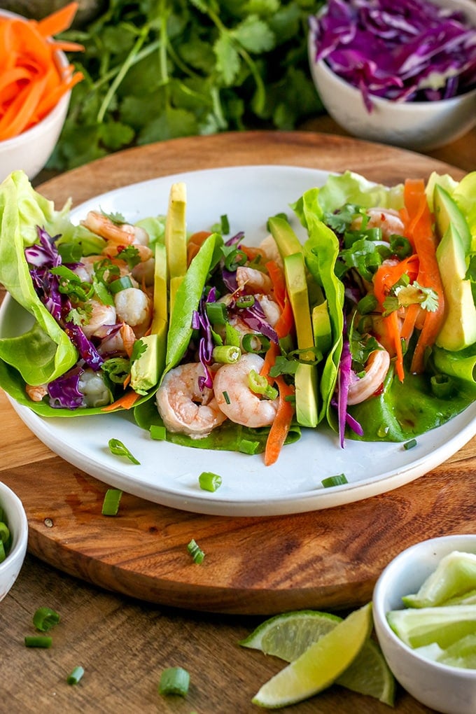 White plate with three lettuce wraps filled with shrimp, red cabbage, cilantro, avocado, green onions and shredded carrots. The plate is sitting on a round wooden cutting board that is surrounded by small bowls filled with chopped green onions, shredded carrots, shredded red cabbage and lime wedges. There is also a bunch of cilantro and half an avocado behind the cutting board.