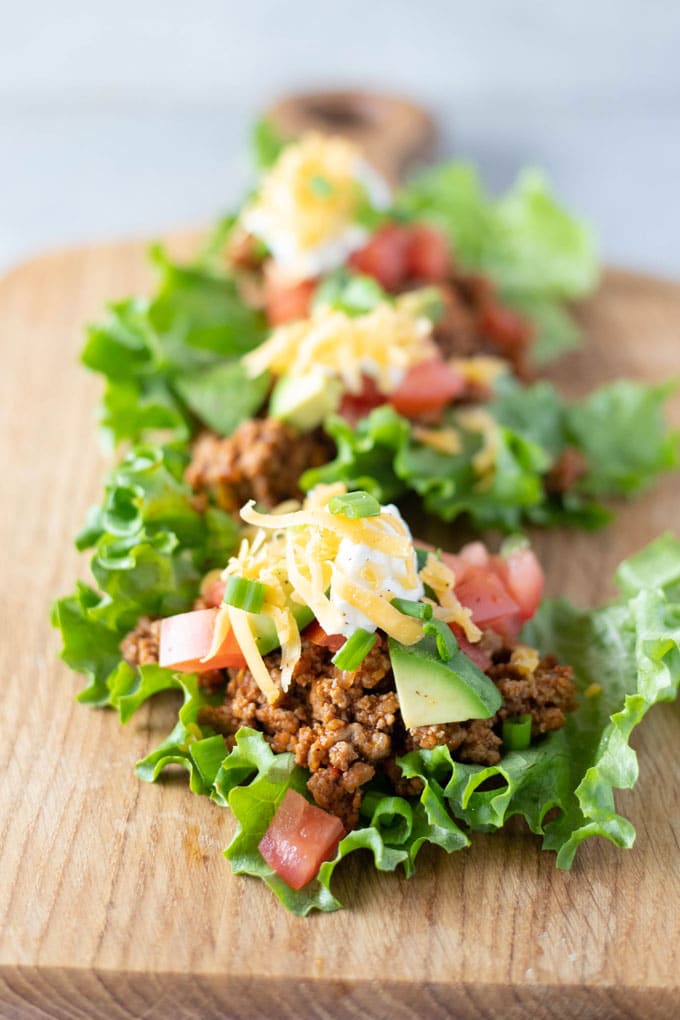 Taco lettuce wraps sitting on a wooden board filled with ground meat, diced tomatoes and avocados, topped with diced green onion, a dollop of sour cream and shredded yellow cheddar cheese.