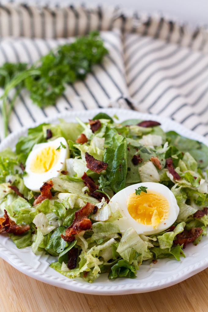 Round white plate filled with chopped lettuce and bacon crumbles, topped with a hard boiled egg that has been halved. The plate is sitting on a blue and white dish towel with a bunch of parsley in the background.