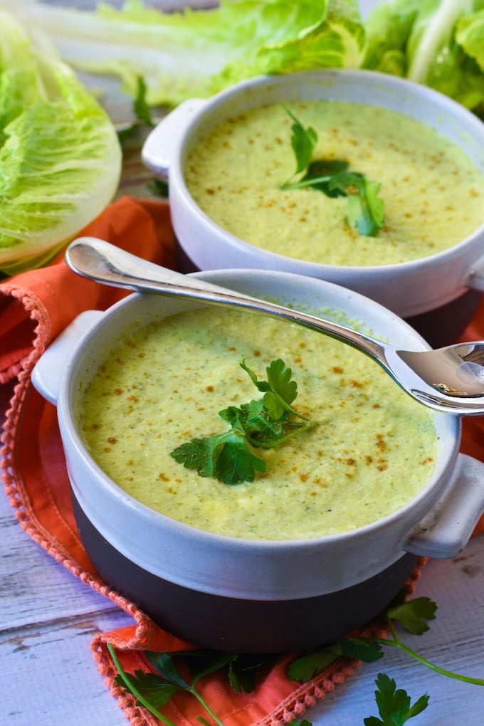 Two white soup bowls filled with light green lettuce soup and garnished with a sprig of parsley. A spoon is sitting across the top of one of the bowls. The bowls are sitting a red napkin and there are leaves of romaine lettuce in the background.