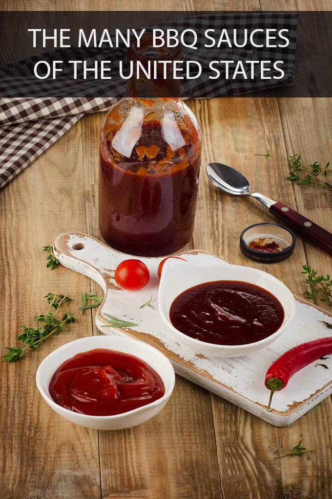 Glass bottle of bbq sauce, half empty, on a wooden table with the bottles lid and a spoon and sprig of thyme to the bottle's right. In front of the bottle is a white cutting board with a white bowl filled with bbq sauce, 2 cherry tomatoes and a red hot pepper. In front of the cutting board is a white bowl full of ketchup, to the left of that is a sprig of thyme. The writing at the top of the picture reads, "The Many BBQ Sauces of the United States"