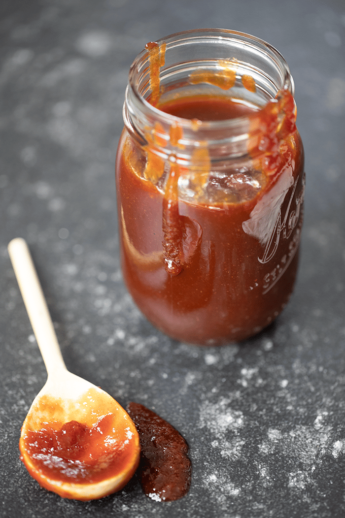 Glass Mason jar, filled almost to the top with bbq sauce, with sauce spilled on the outside of the jar. Jar is sitting on a dark grey surface, and there is a white plastic spoon that had been dipped in the sauce in front of the jar.