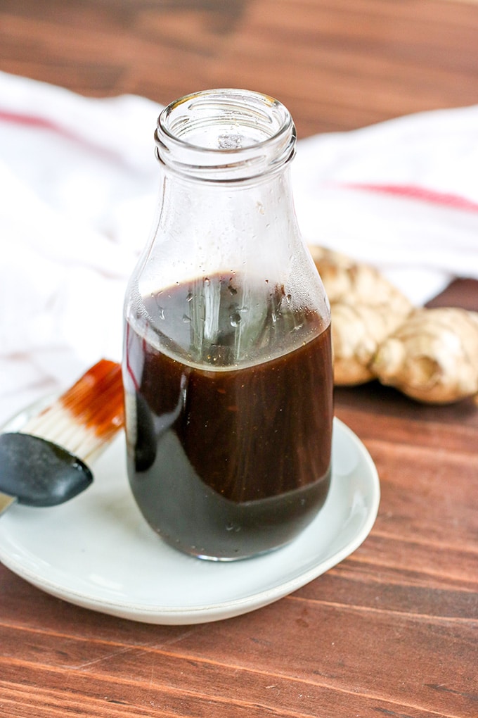 Glass milk bottle ¾ filled with brown Korean bbq sauce on a small white plate. A brush that has been dipped in the sauce is resting on the place and a knob of ginger and dishtowel are in the background