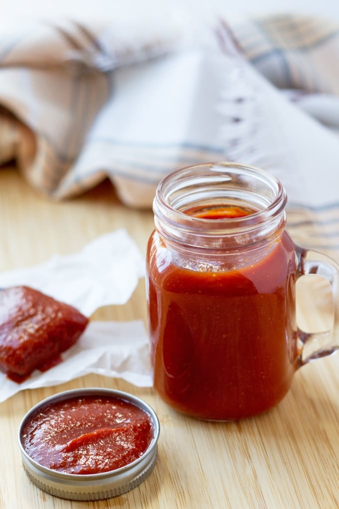 Glass mason jar mug of bbq sauce on a wooden table with the lid, full of sauce, next to it and a brush, wet with sauce, on a piece of parchment paper behind it.