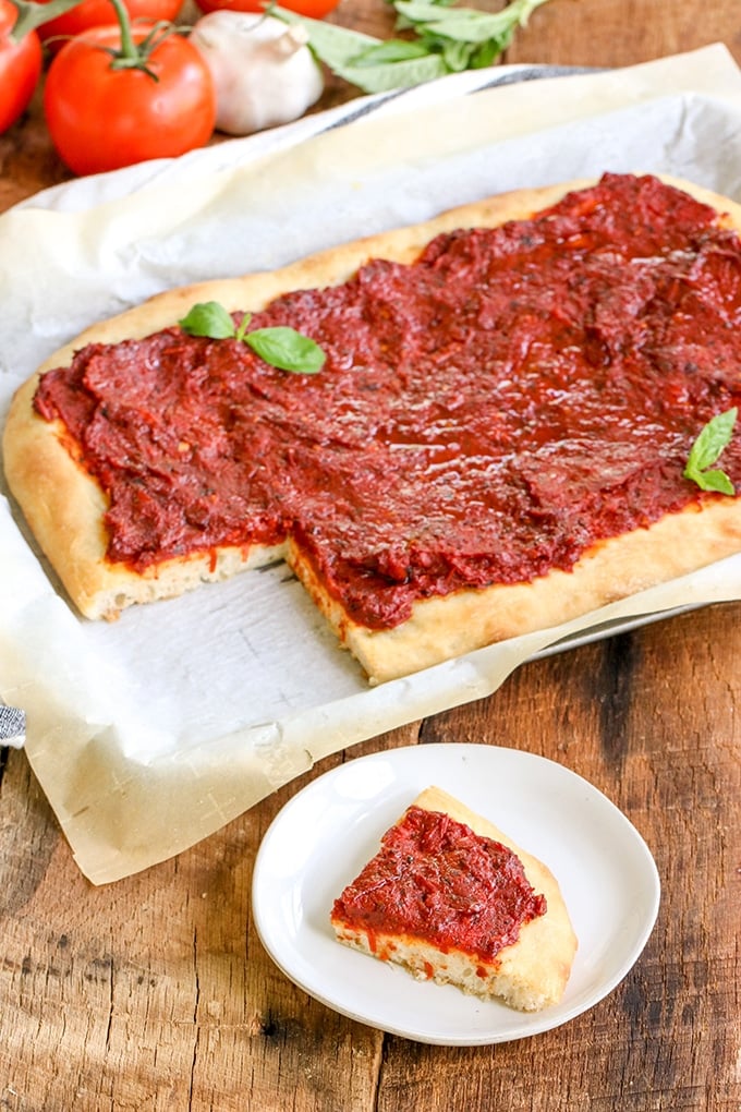 Italian Tomato Pie on a parchment paper rimmed baking sheet sitting on a wooden surface; in the background is a bulb of fresh garlic, some fresh basil leaves and some tomatoes on the vine.