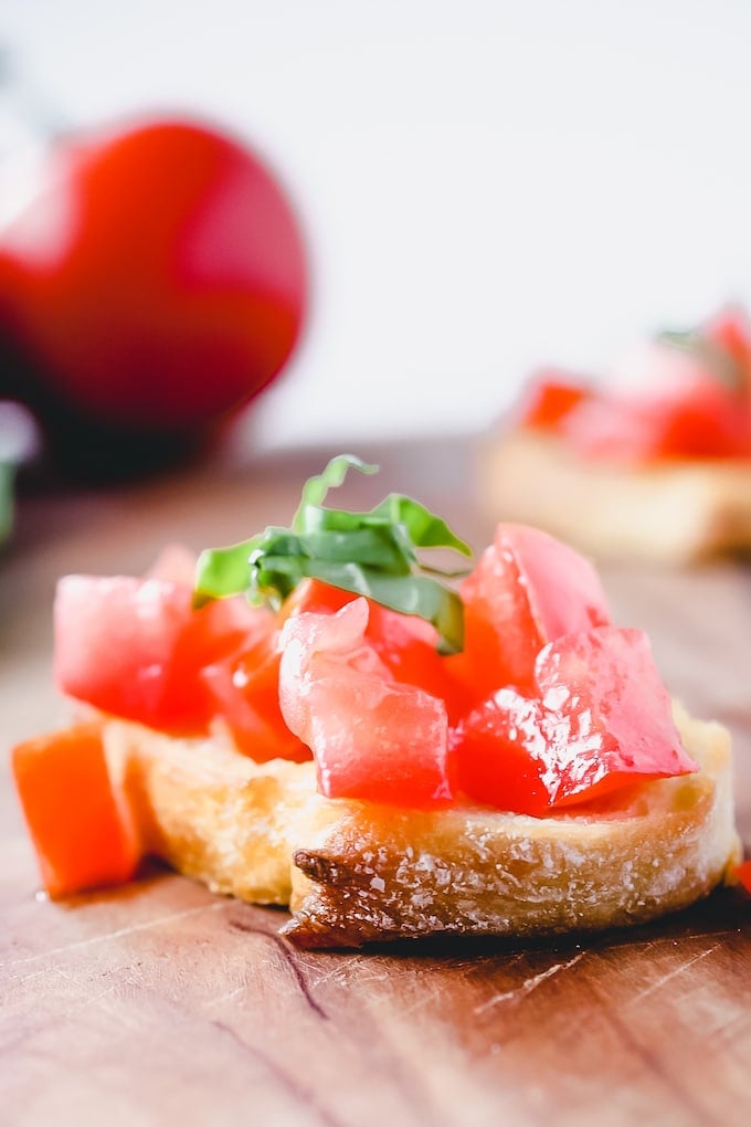 A close up picture of a toasted sliced of baguette topped with chopped tomatoes and garnished with ribbons of chopped fresh basil; in the background you can see a whole tomato.