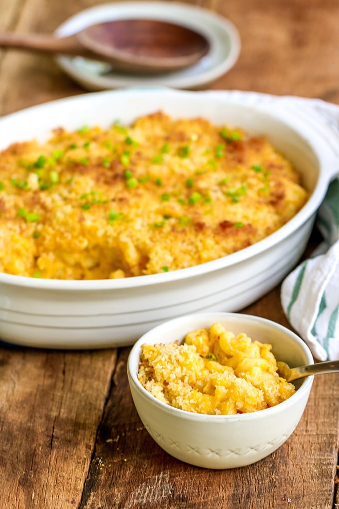 White oval ceramic baking dish of golden baked mac and cheese topped with breadcrumbs and chopped chives with a scoop taken out of it. A small white bowl filled with mac and cheese sits in front of the baking dish with a spoon in it.