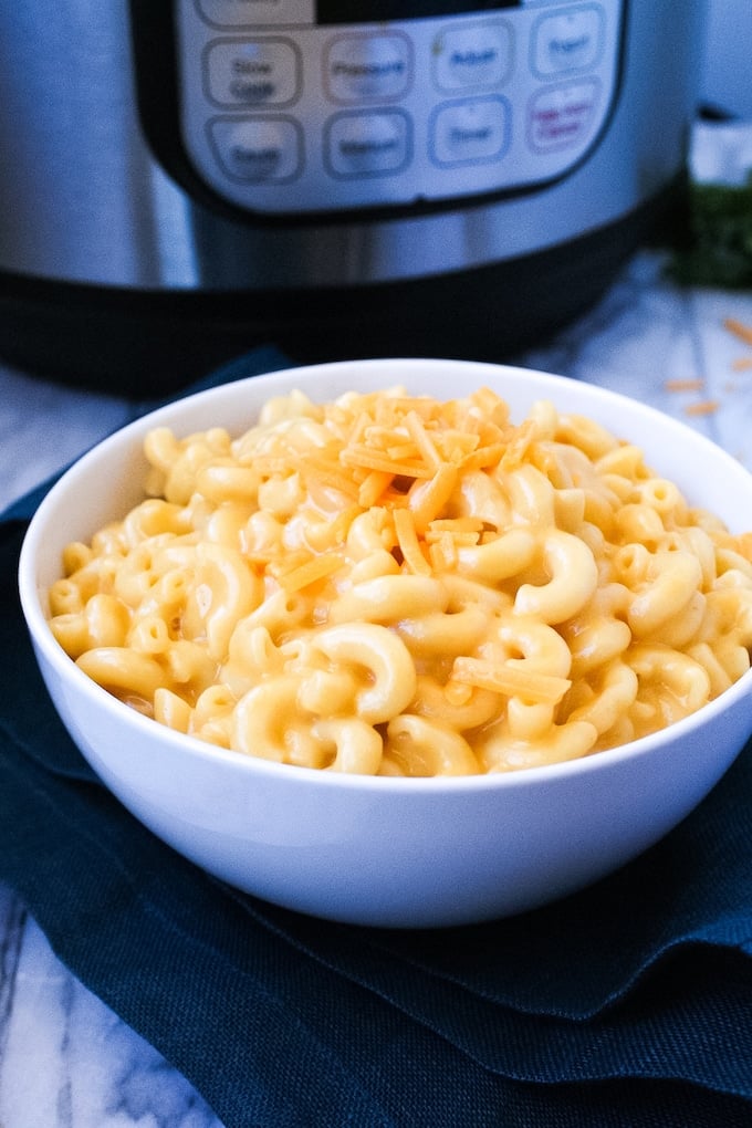 White bowl sitting in front of an instant pot filled with cream macaroni and cheese topped with shredded cheddar cheese.
