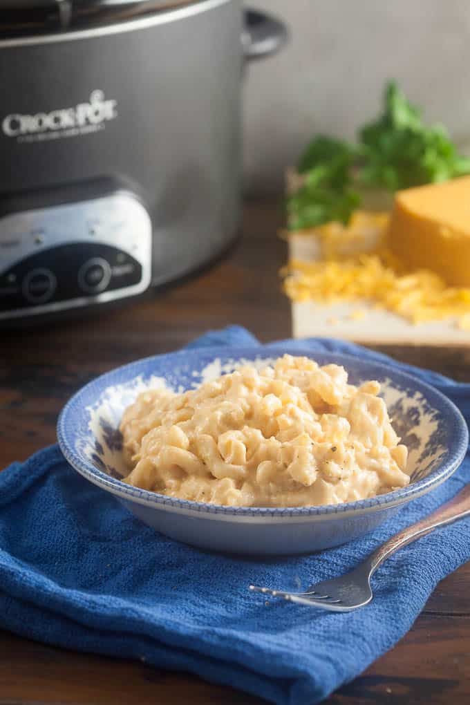 Blue and white bowl filled with creamy mac and cheese sitting on a blue cloth napkin with a fork on it. In the background is a slow cooker, and a block of yellow cheddar cheese that has been partially shredded and a some parsley.