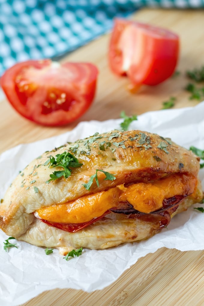 Baked chicken breast stuffed with yellow cheddar cheese, deli meat and tomatoes sitting on a wrinkled piece of white parchment paper sprinkled with chopped parsley; the parchment paper and chicken breast are sitting on a wooden cutting board; in the background is a halved tomato.