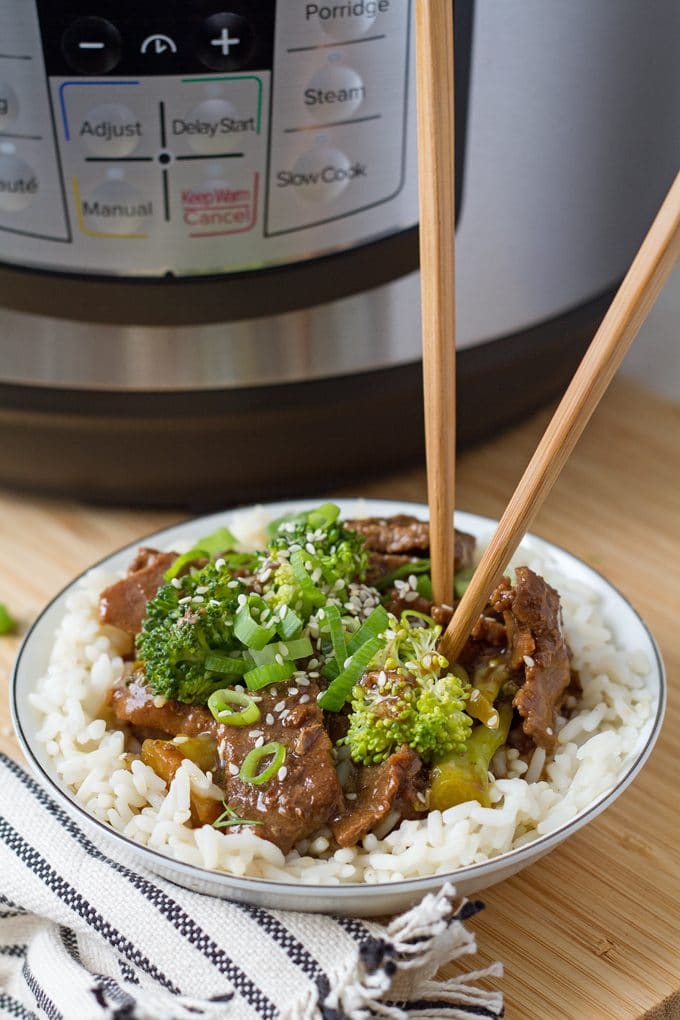 White bowl filled with white rice and topped with beef with broccoli. It has been garnished with green onions and sesame seeds. The bowl has two wooden chopsticks sticking out of it, and is sitting on a wooden surface in front of a slow cooker. There is a white and blue stripped cloth napkin under the bowl.