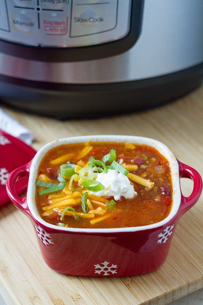 Small red, square ceramic dish with a white snowflake pattern and handles is filled with chili topped with shredded cheddar cheese, green onions and a dollop of sour cream. It is sitting on a wooden surface in front of an Instant Pot.