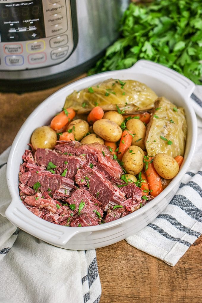 White ceramic oval shaped dish filled with corned beef, small white potatoes and baby carrots, and cabbage that has been sprinkled with chopped parsley. In the background is an instant pot and a bunch of parsley. The dish is sitting on a white and blue stripped dish towel.