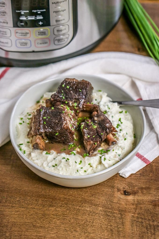 White bowl on a brown wood surface filled with mashed potatoes, topped with three beef short ribs and topped with finely chopped chives. The bowl is sitting in front of an instant pot. Next to the instant pot is a bunch of chives. Between the instant pot and bowl is a white dishtowel with a red stripe.