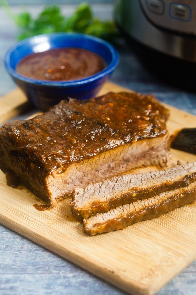 Piece of brisket sitting on a wooden cutting board, covered in BBQ sauce, that has had two slices cut from it. A small blue bowl of bbq sauce is behind it on the cutting board. In teh background is an Instant Pot.