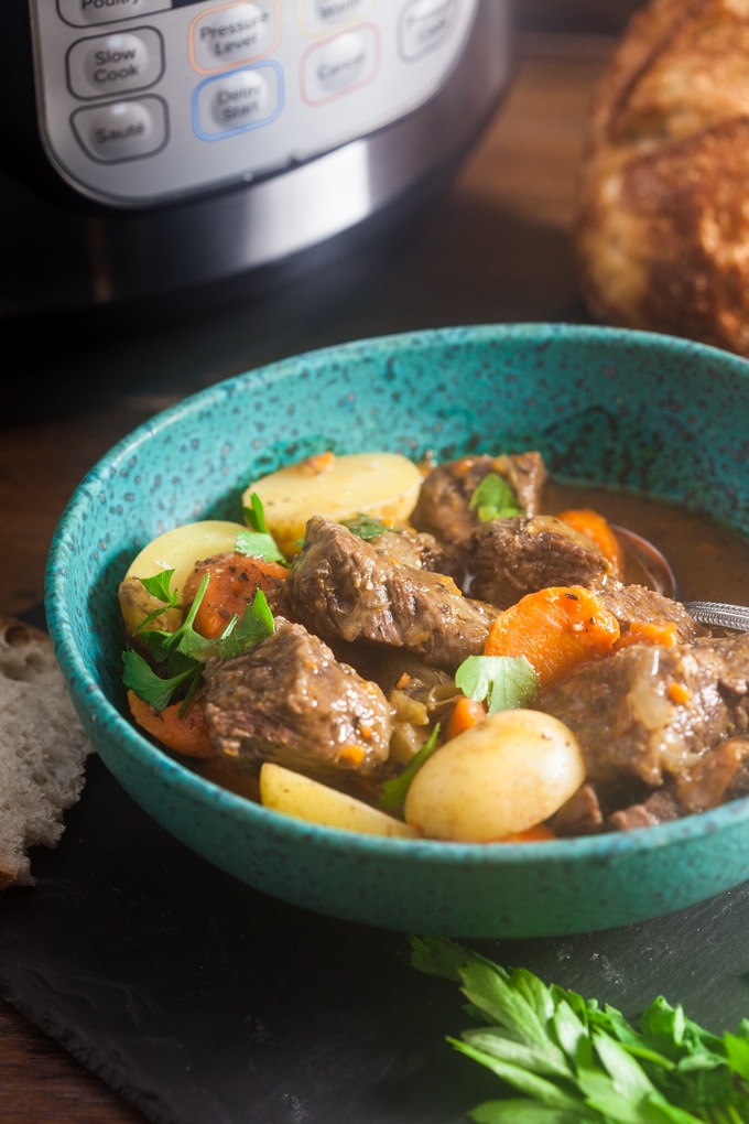 Speckled blue bowl filled with stewed beef chunks, carrots and potatoes topped with chopped parsley. There is a spoon in the bowl. The bowl in in front of the instant pot and a loaf of crusty bread.
