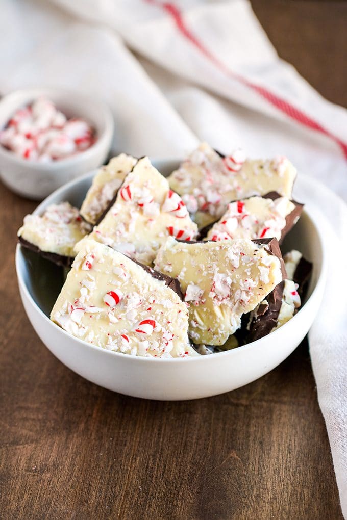 white bowl filled with broken pieces of peppermint bark (milk chocolate, with a layer of white chocolate then topped with crushed peppermint candies); the bowl is on a wooden surface; to the right of the bowl is a dish towel with a red stripe and behind the bowl is a small white bowl with whole peppermint candies.