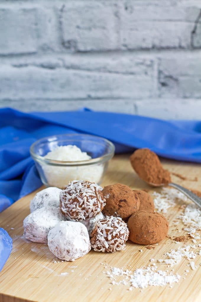 rum balls rolled in three different toppings (powdered sugar, coconut and cocoa powder) sitting on a wooden cutting board; cutting board is sprinkled with powdered sugar, cocoa and coconut; small glass bowl of coconut is sitting at the corner of the cutting board behind the rum balls; to the right of the rum balls on the cutting board is a metal spoon full of cocoa powder; blue cloth napkin in the background.