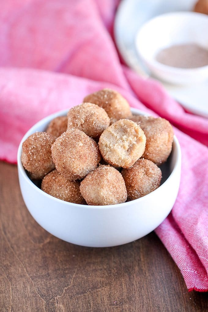 white bowl filled with snickerdoodle bites covered in cinnamon sugar; bowl is sitting on a wooden surface; pink dishtowel is behind the bowl; behind dishtowel is a white plate with a small bowl of cinnamon sugar.