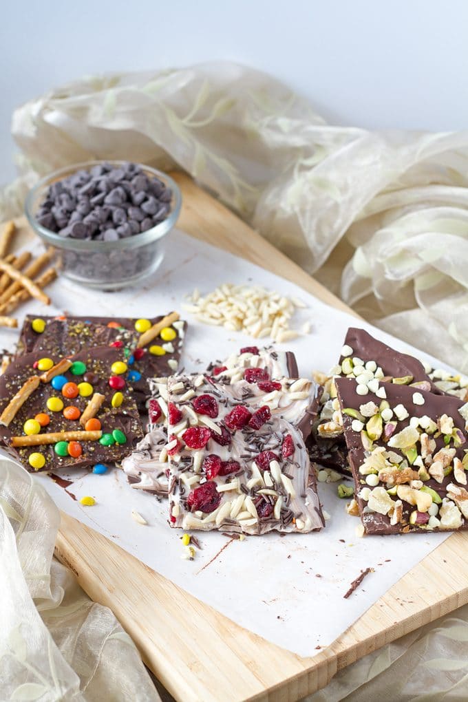 three different stacks of pieces of chocolate barks (topped with M & Ms and pretzels, topped with chocolate sprinkles, almond pieces and dried cranberries and topped with mixed nuts) sitting on a piece of parchment paper on a wood cutting board; a glass bowl of chocolate chips is sitting at one end of the board next to   a stack of pretzel sticks; board is surrounded by flowy, light yellow fabric.