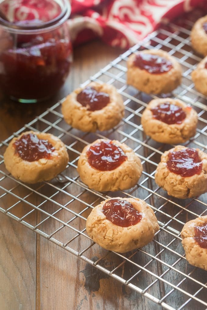 no-bake peanut butter thumb print cookies filled with strawberry jam sitting on a wire rack on a wooden surface; to the left at of the rack of cookies is a glass jar of strawberry jam which is in front of a red and white dish cloth.