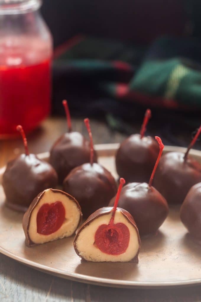 cherry peanut butter balls (with the cherry stems sticking out) on round  wooden plate; one is cut in half so you can see the cherry inside; jar of maraschino cherries and a green dishtowel in the background.