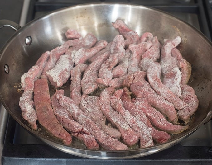 Skillet of raw beef strips tossed in flour.