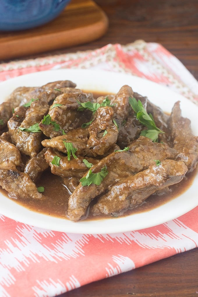 Beef strips in gravy on a white plate sprinkled with chopped parsley. The white plate is on a red and white placemat and in the background you can see the corner of a wood cutting board and the bottom of blue pot.