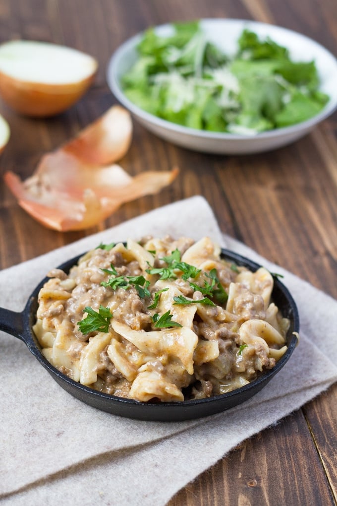 Small cast-iron skillet filled with beef stroganoff garnished with parsley. The skillet is on top of a piece of tan felt. In the background on the wood table is a some onion skin, a halved onion and a white bowl full of salad.