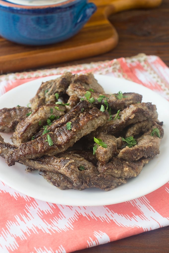 Browned beef strips on a white plate sprinkled with chopped parsley. The plate is on a red and white placemat. Behind the plate is a wooden cutting board with a blue bowl on it.