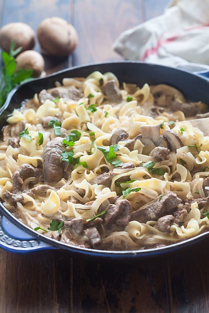 Blue skillet sitting on a wood table filled with beef stroganoff with 3 mushrooms, some parsley and a dish towel in the background.