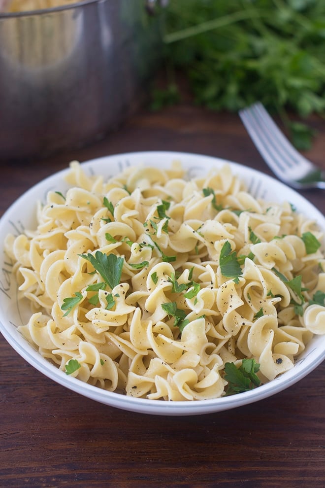 White bowl filled with egg noodles garnished with chopped parsley and black pepper. The bowl sits on a wood table and there is fork and a bunch of flat leaf parsley in the background.