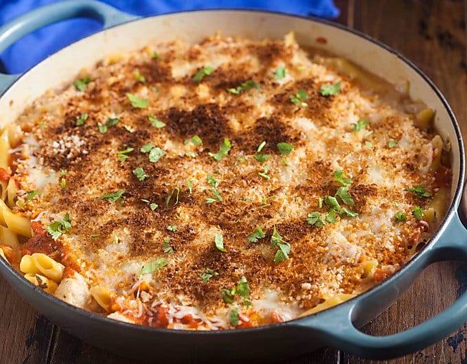 shallow blue pan filled with Chicken Parmesan pasta bake; pasta is tapped with melted white cheese topped with golden bread crumbs and garnished with chopped parsley; pot is sitting a a wooden surface; you can see a blue cloth napkin in the background.