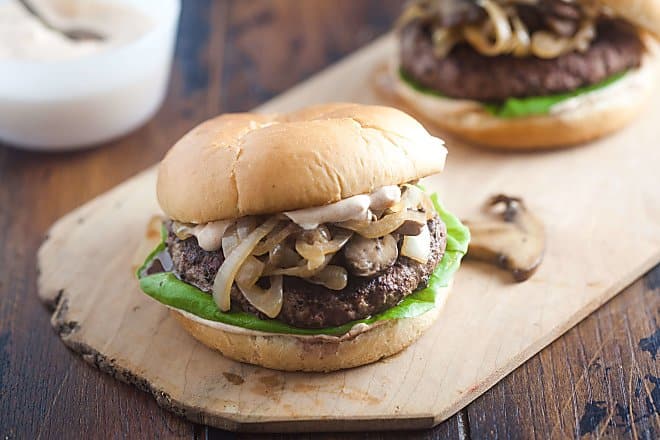 Beef Stroganoff Burger with sautéed mushrooms and onions, lettuce and white sauce sitting on a thin wooden cutting board with a slice of mushroom in the background as well as another burger. The thin wooden cutting board is sitting on a wooden table and there is a jar of sauce in the background.