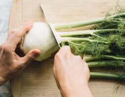 A fennel bulb on a cutting board. The stalks and fronds are being cut away from the bulb.